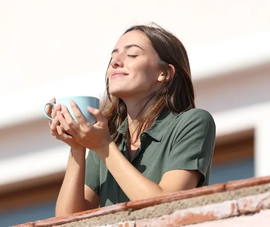 Woman on a balcony holding a blue mug, eyes closed in the sun